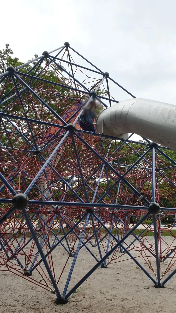 A child climbing the large blue-and-red rope pyramid beside the tube slide at West Coast Park.